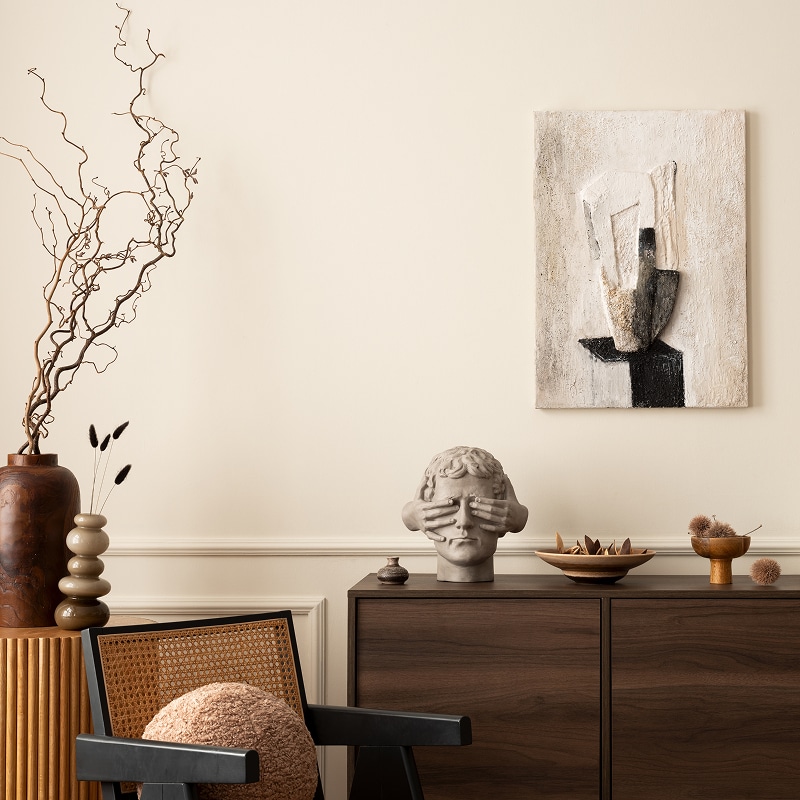 Décoration moderne: mur beige, sideboard bois foncé, fauteuil rotin noir. Sculpture de tête avec mains couvrant les yeux et art abstrait texturé.